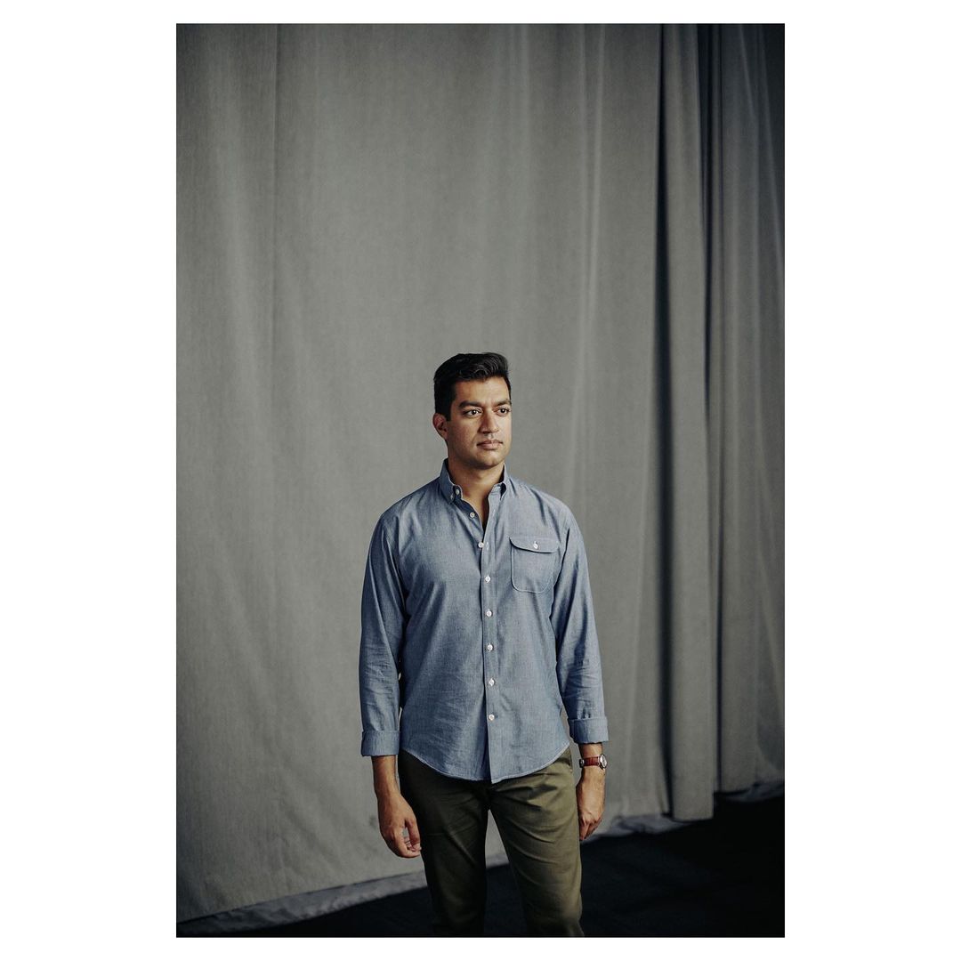 Man wearing a blue button-down shirt and dark trousers standing before a grey draped curtain backdrop.