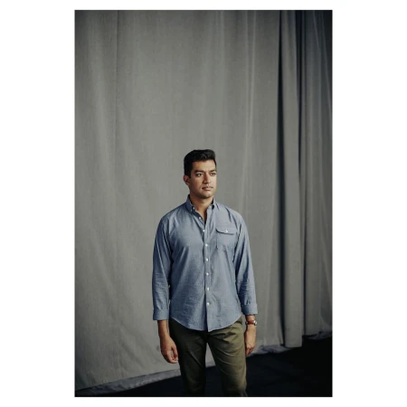 Man wearing a blue button-down shirt and dark trousers standing before a grey draped curtain backdrop.