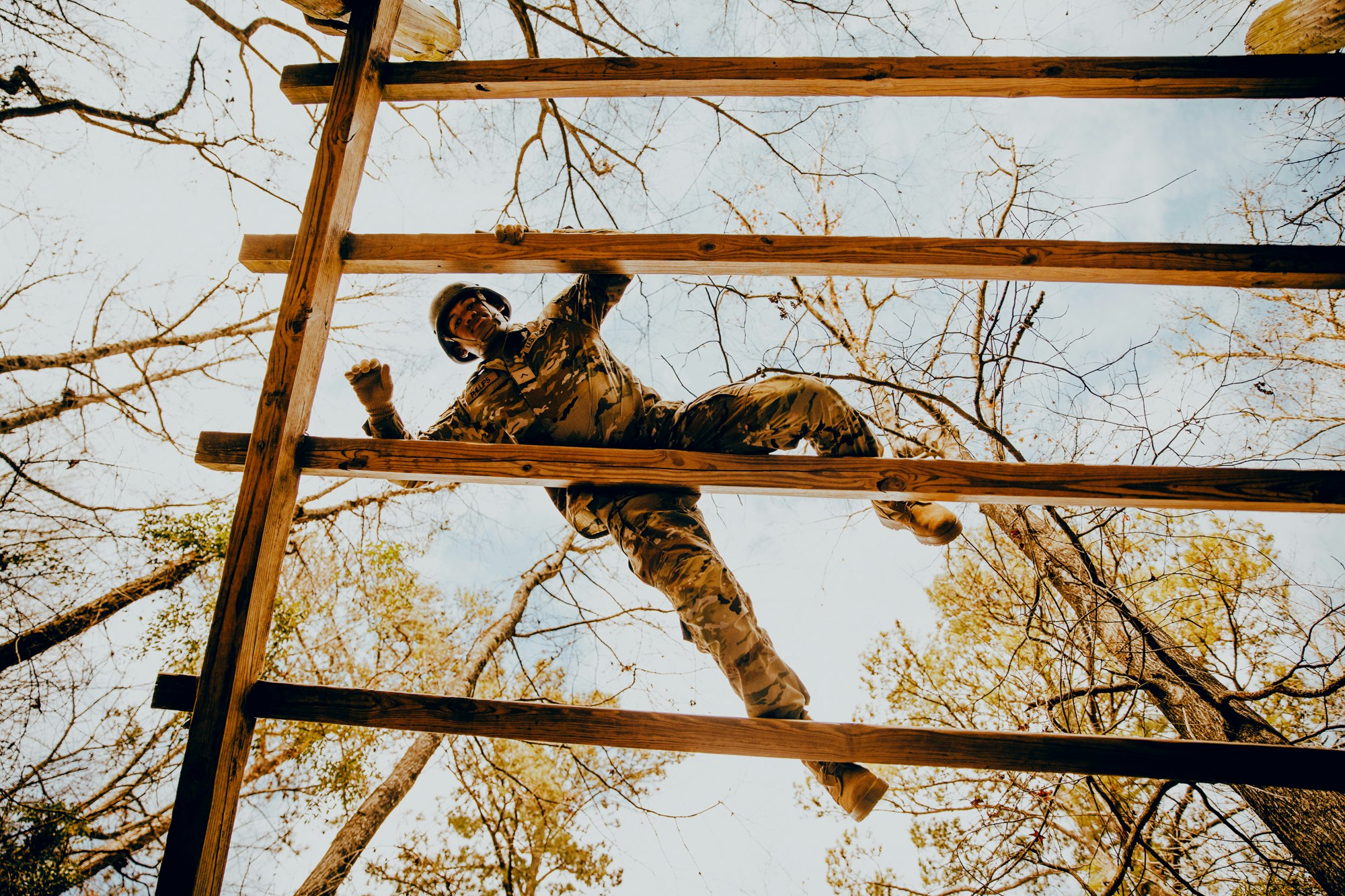 Man in camouflage climbing wooden ladder in forest. Low angle view of obstacle course, trees, and sky. Outdoor activity, fitness, and determination.