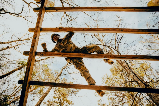 Man in camouflage climbing wooden ladder in forest. Low angle view of obstacle course, trees, and sky. Outdoor activity, fitness, and determination.