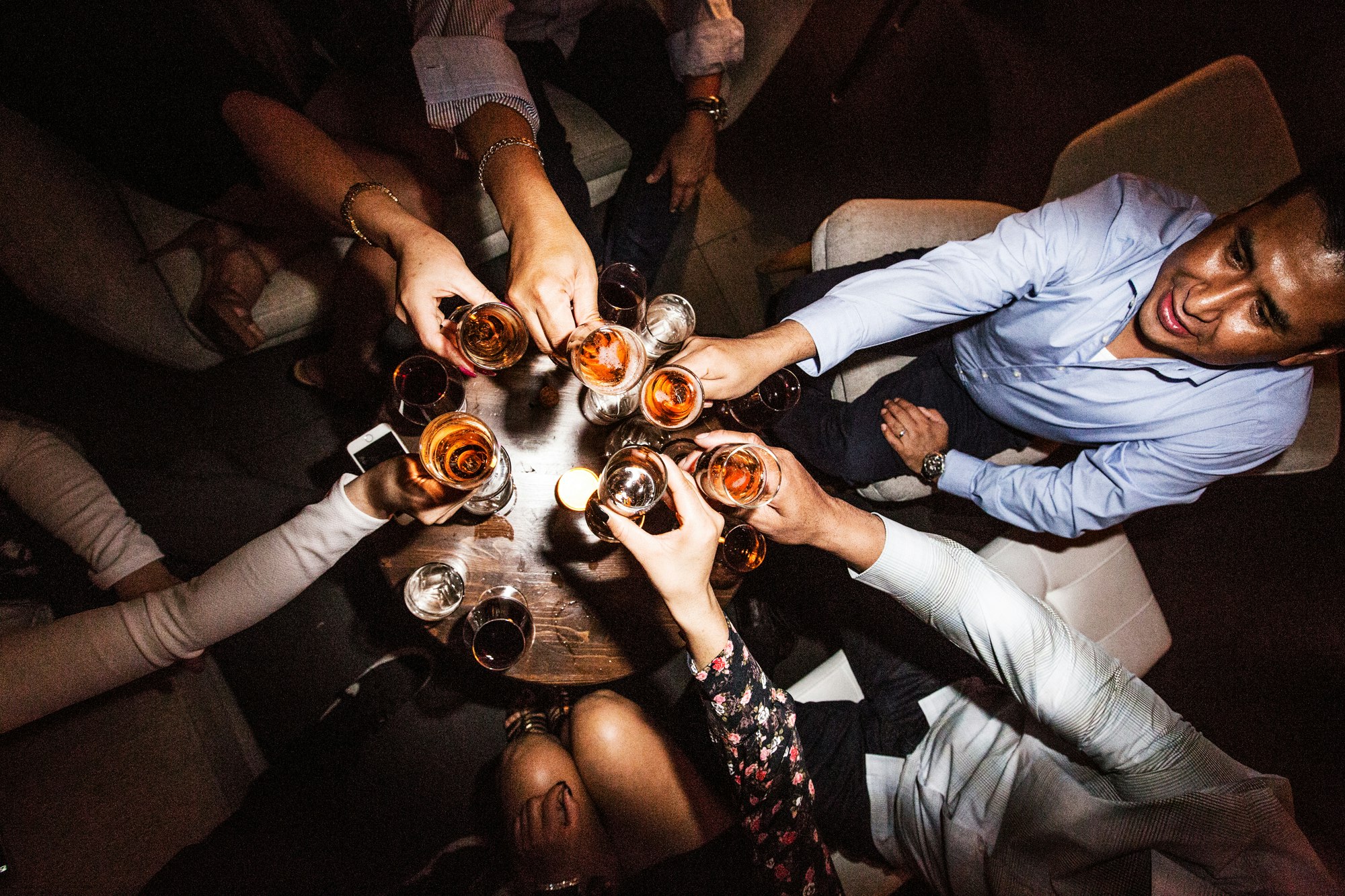 Overhead shot of diverse people toasting with drinks at a wooden table in a dimly lit indoor bar or lounge environment. Cheers!
