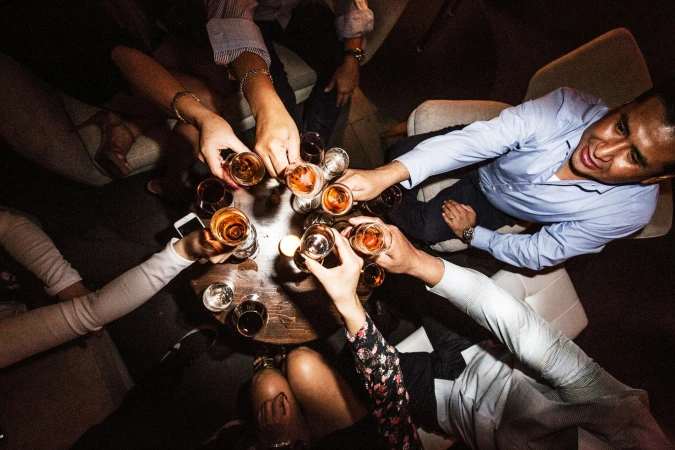 Overhead shot of diverse people toasting with drinks at a wooden table in a dimly lit indoor bar or lounge environment. Cheers!