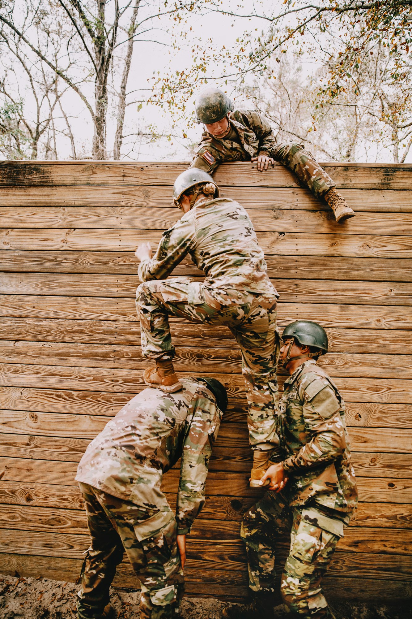 Four military adults work together to scale a wooden wall in an outdoor training environment, exhibiting teamwork and physical skill.