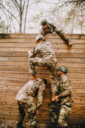 Four military adults work together to scale a wooden wall in an outdoor training environment, exhibiting teamwork and physical skill.