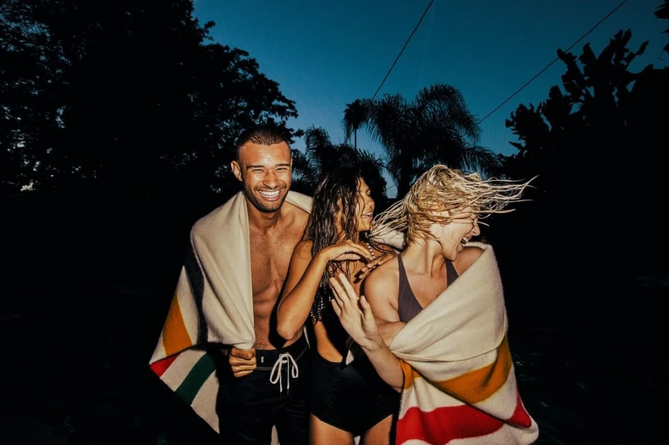 Three people wrapped in blankets smile at the camera at night near trees. One man, two women, outdoor setting, dark mood.
