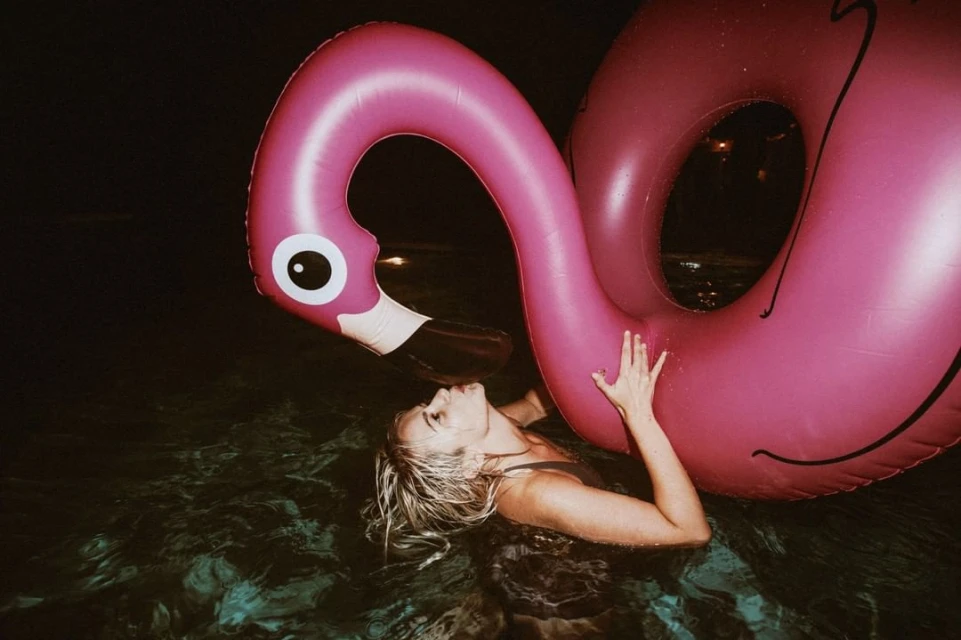 A woman swimming in a pool at night, partially submerged and holding a large pink flamingo inflatable pool toy for flotation.