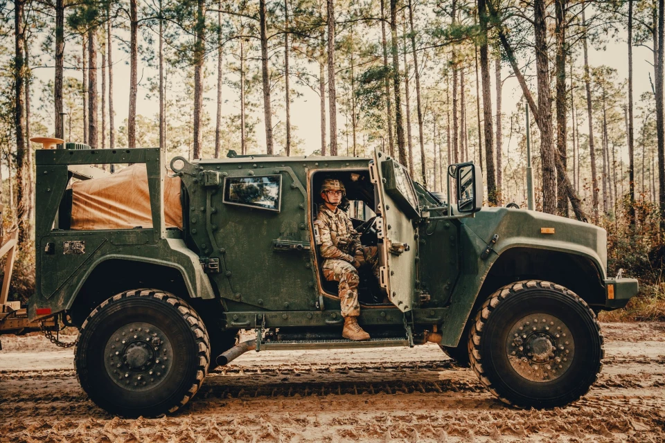 Military vehicle with a soldier in camouflage uniform driving through a forest of tall trees on a dirt road in an outdoor setting.
