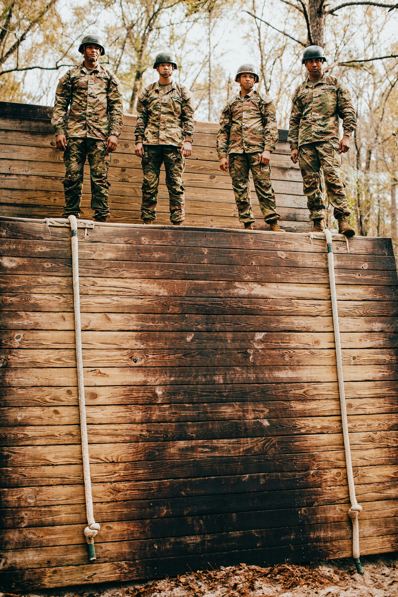 Four uniformed soldiers stand on a wooden wall outdoors against a background of trees, showcasing a display of military teamwork.