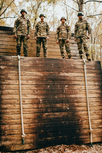 Four uniformed soldiers stand on a wooden wall outdoors against a background of trees, showcasing a display of military teamwork.