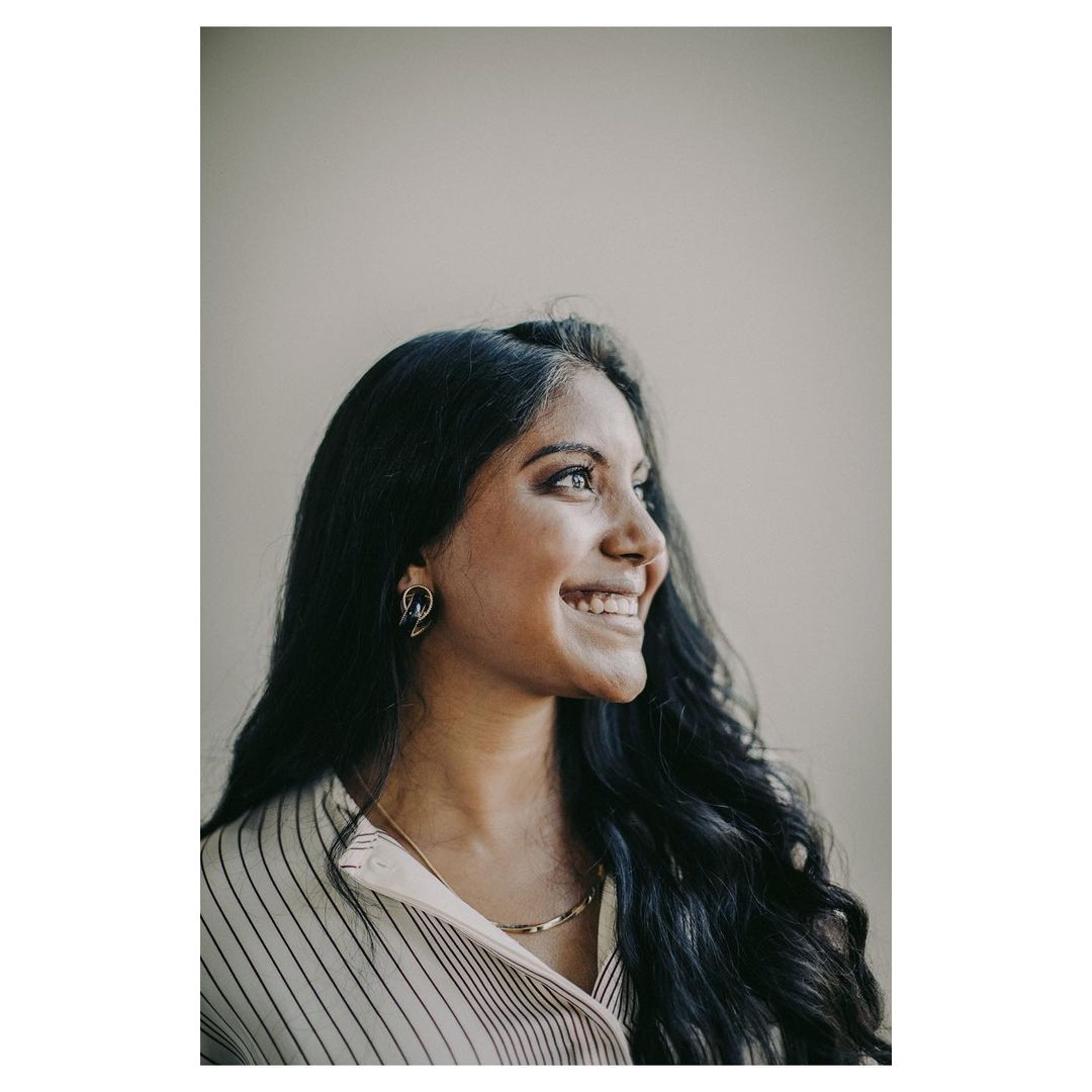 Smiling woman with dark wavy hair, striped shirt, and gold necklace poses indoors against a neutral background in a portrait.