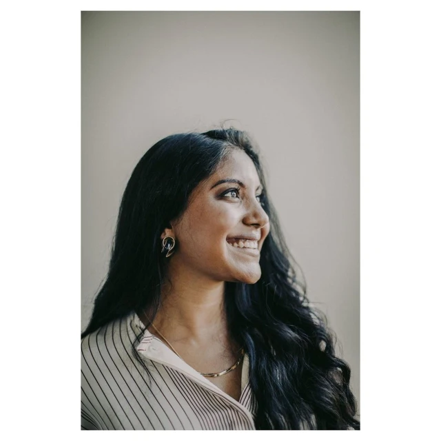 Smiling woman with dark wavy hair, striped shirt, and gold necklace poses indoors against a neutral background in a portrait.