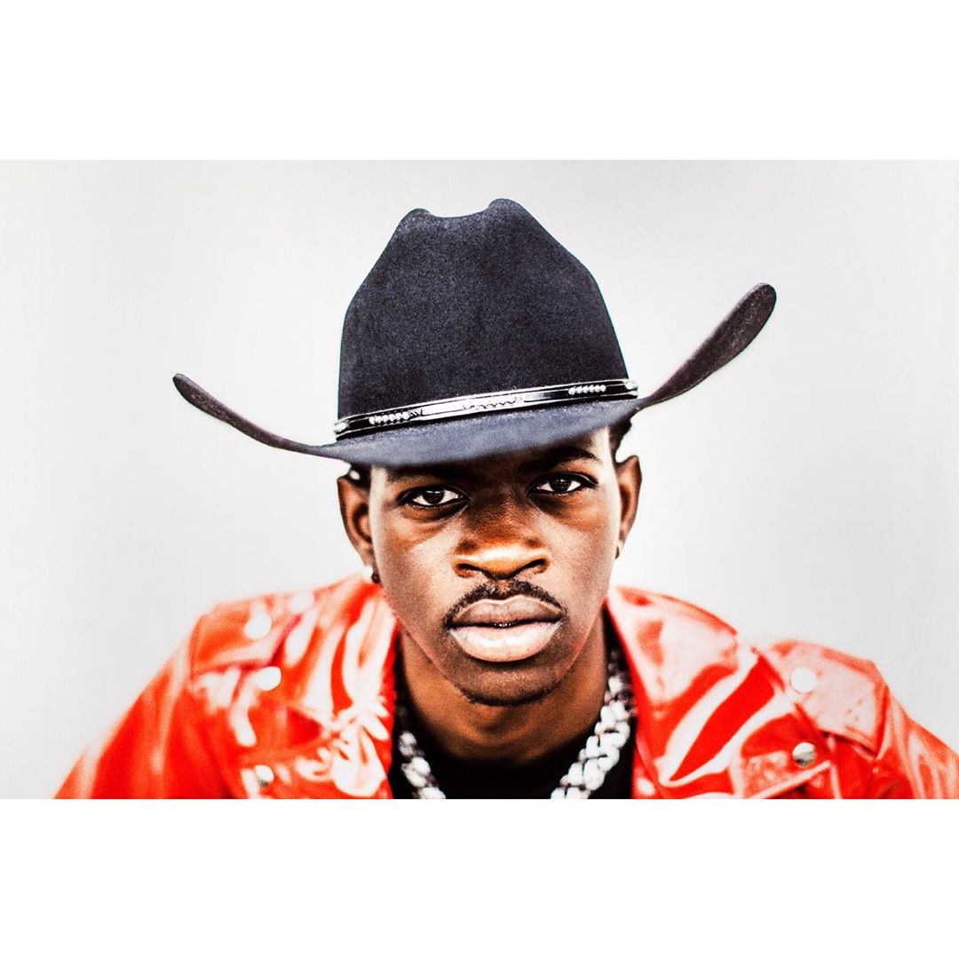 Close-up of Lil Nas X wearing a black cowboy hat, red leather jacket, and white chain against a neutral background.