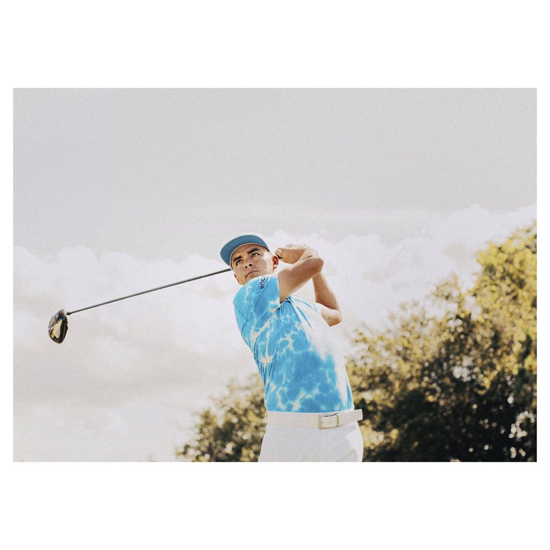 Male golfer in mid-swing on a sunny day, wearing a blue tie-dye shirt and white pants, set against a background of trees and sky.