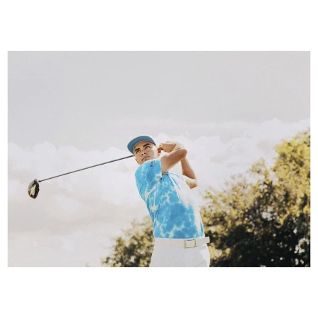 Male golfer in mid-swing on a sunny day, wearing a blue tie-dye shirt and white pants, set against a background of trees and sky.