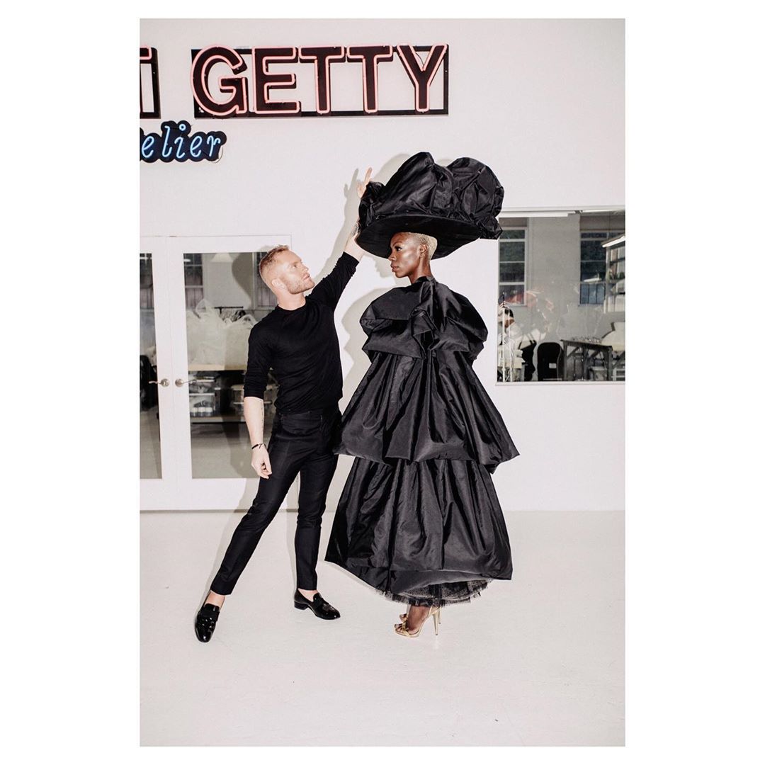 Fashion shoot in photo studio, featuring a stylist adjusting a model's oversized black tiered taffeta gown and wide-brimmed black hat.
