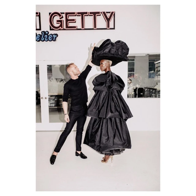 Fashion shoot in photo studio, featuring a stylist adjusting a model's oversized black tiered taffeta gown and wide-brimmed black hat.