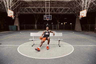 Black man sitting on a silver bench on an outdoor basketball court holding a basketball at night.