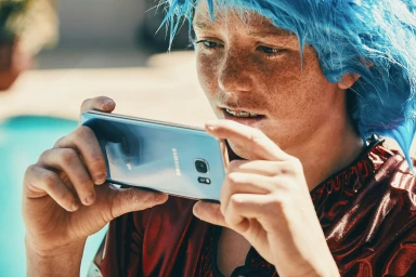 Young person with bright blue hair and freckles holding and photographing with a smartphone outdoors