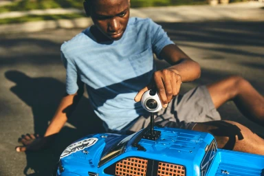Young Black boy sitting on asphalt adjusting a camera mounted on a blue remote control truck