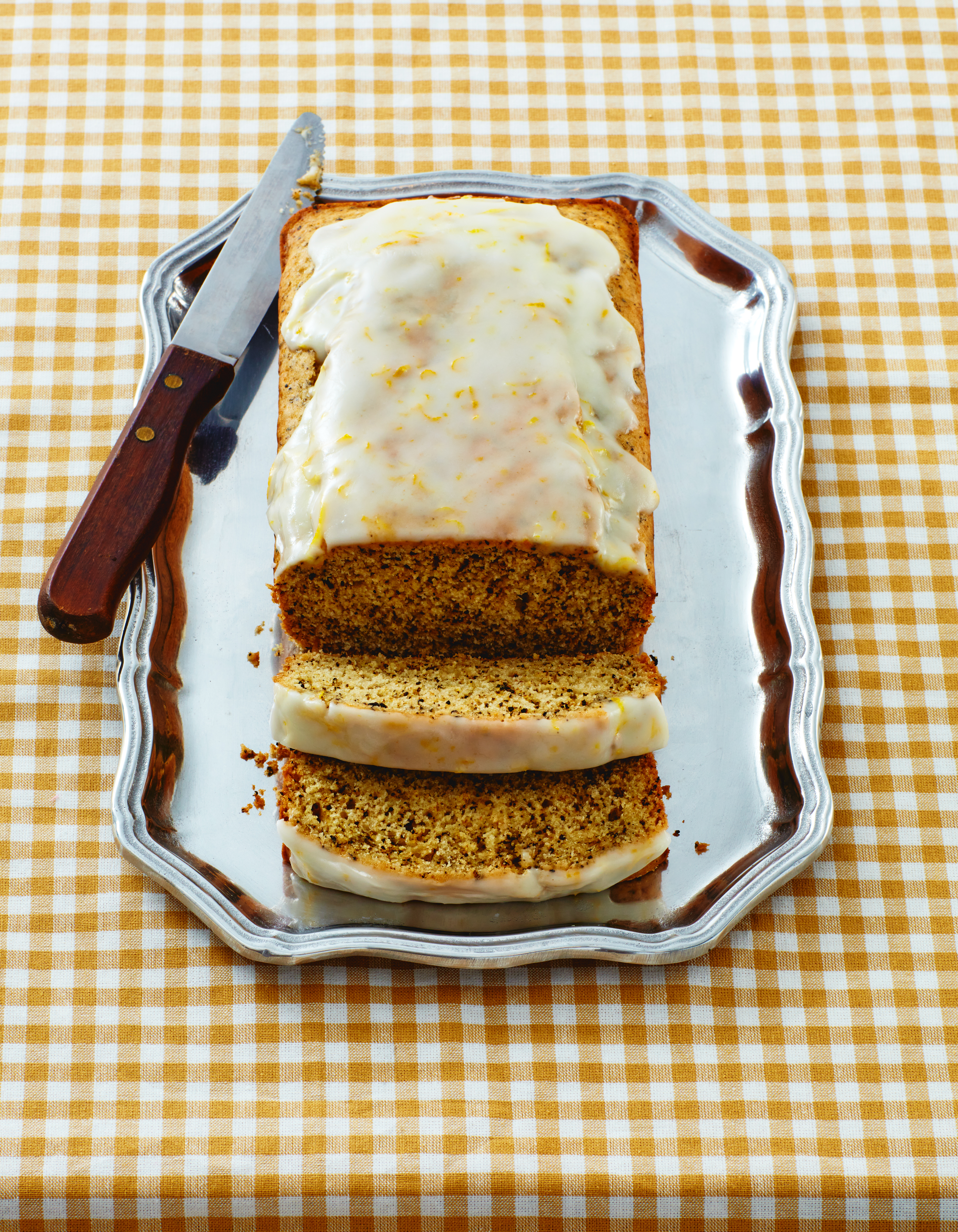 Overhead shot of iced loaf cake sliced on a silver tray and a knife on a yellow and white checkered tablecloth background.