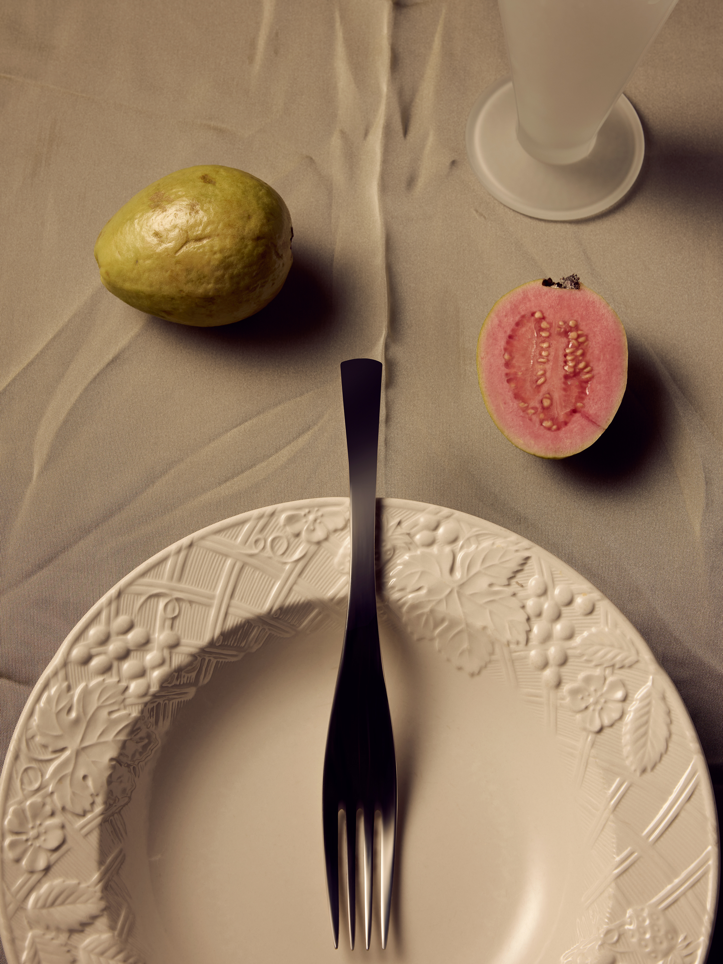 Overhead shot of a table setting with a plate, fork, guava fruit, and a clear glass on a textured, neutral-toned cloth background.