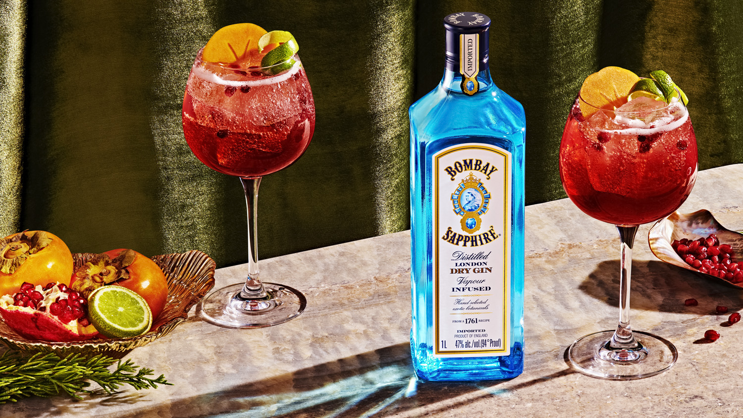 Bombay Sapphire gin bottle, two cocktails with fruit garnish, and fresh fruit on a textured surface with a draped olive-green background.
