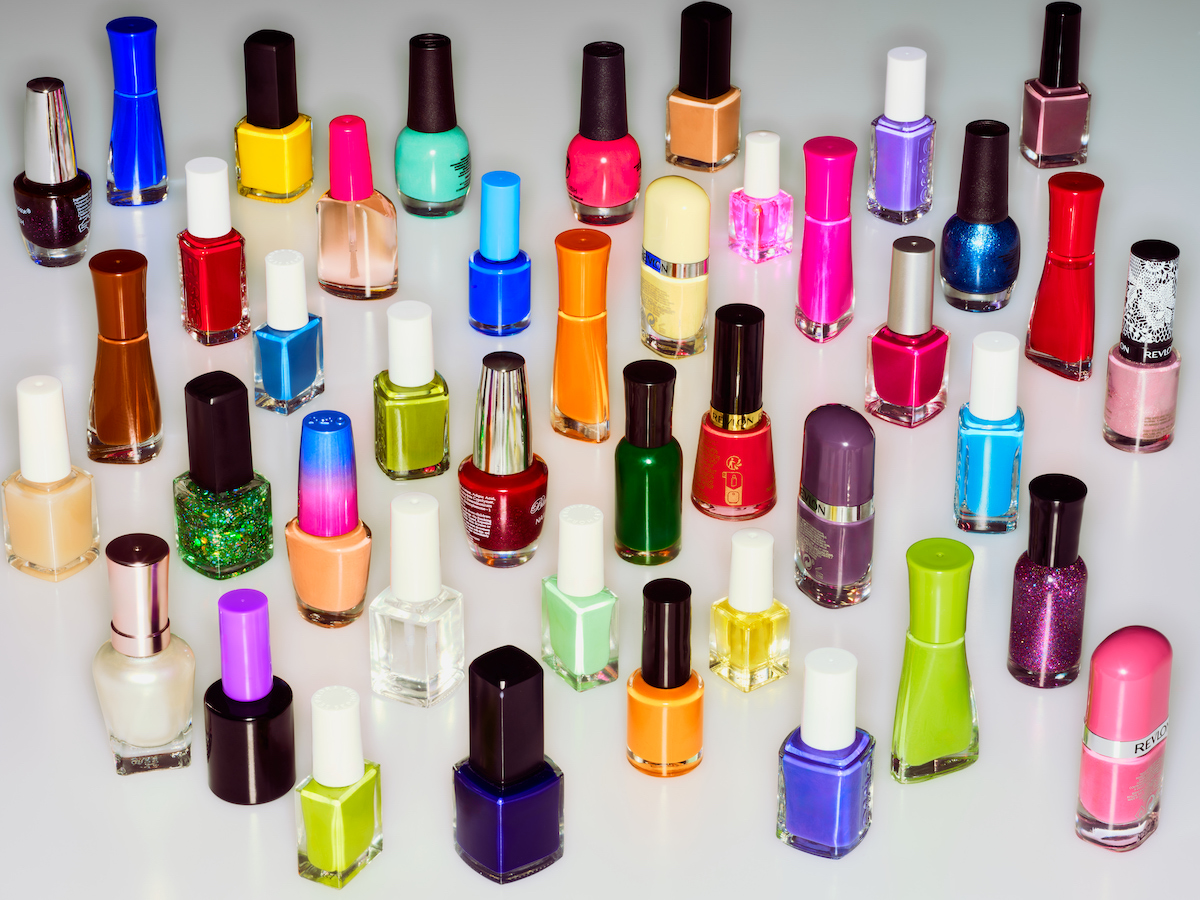 Overhead shot of assorted colorful nail polish bottles with black caps, neatly arranged on a white background, providing a beauty product display.
