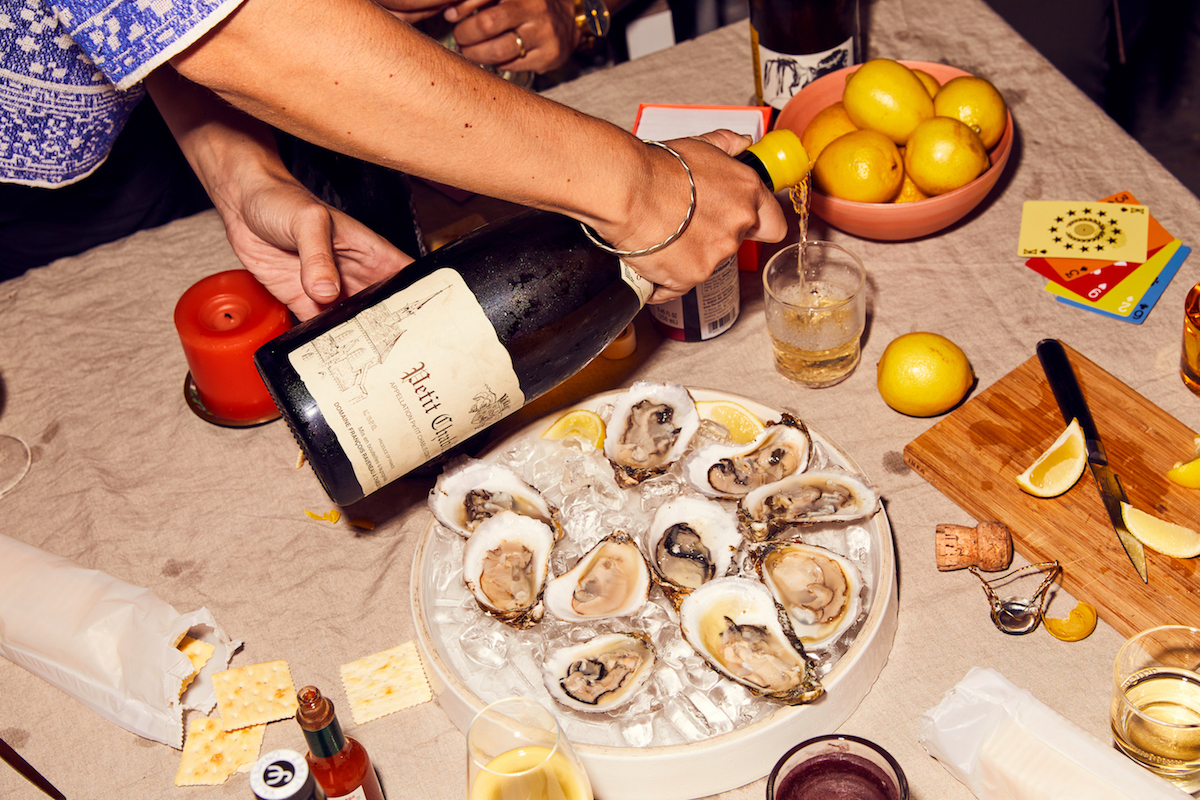Overhead shot of an oyster platter, wine bottle being poured into a glass, lemons, crackers, and sauce on a linen-covered table.