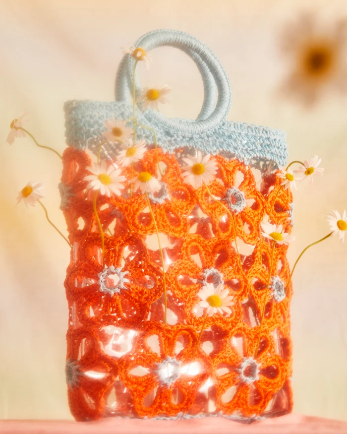 Orange floral crochet handbag with pale blue circular handles filled with fresh white daisies in a warm diffused setting.