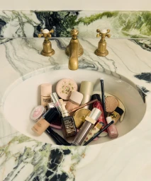 Collection of cosmetic products and makeup brushes arranged inside a white bathroom sink with green marble countertops and brass faucets.