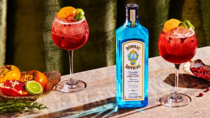 A bottle of Bombay Sapphire gin flanked by two cocktail glasses filled with a red drink, garnished with citrus slices on a marble surface.