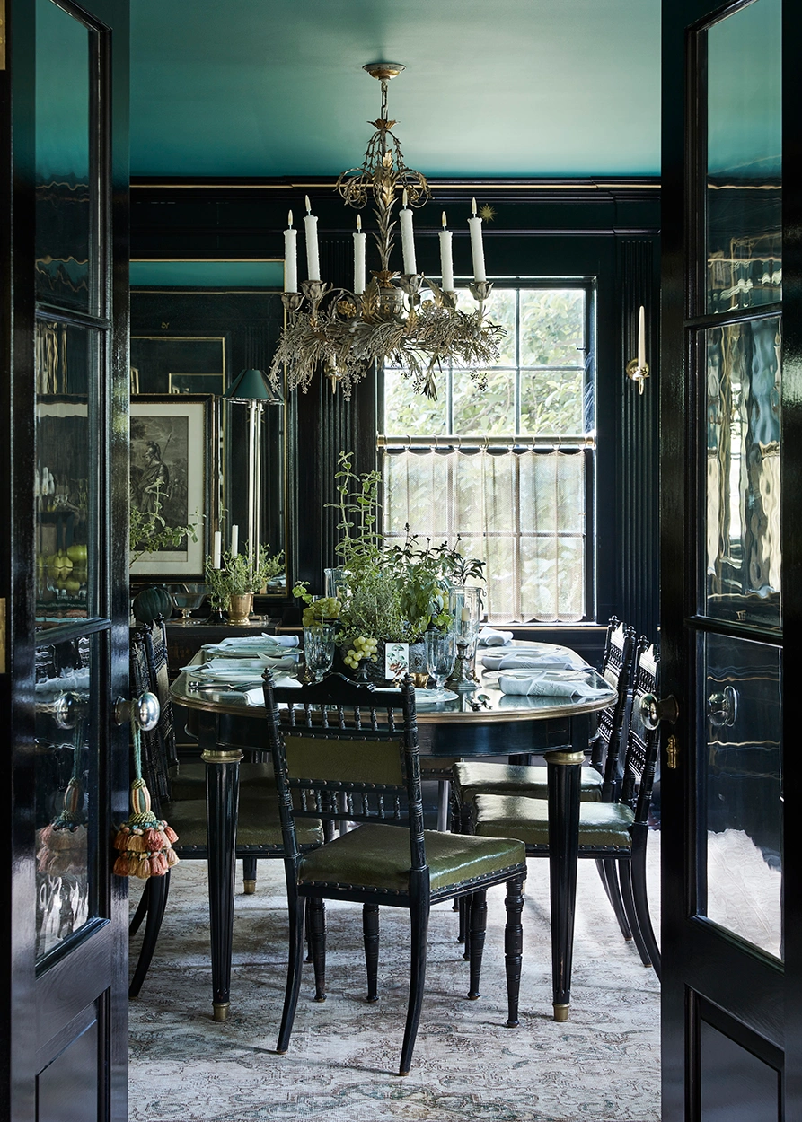 View through dark double doors into an elegant dining room styled by Sophie Strangio with a dark table, chandelier, and green accents