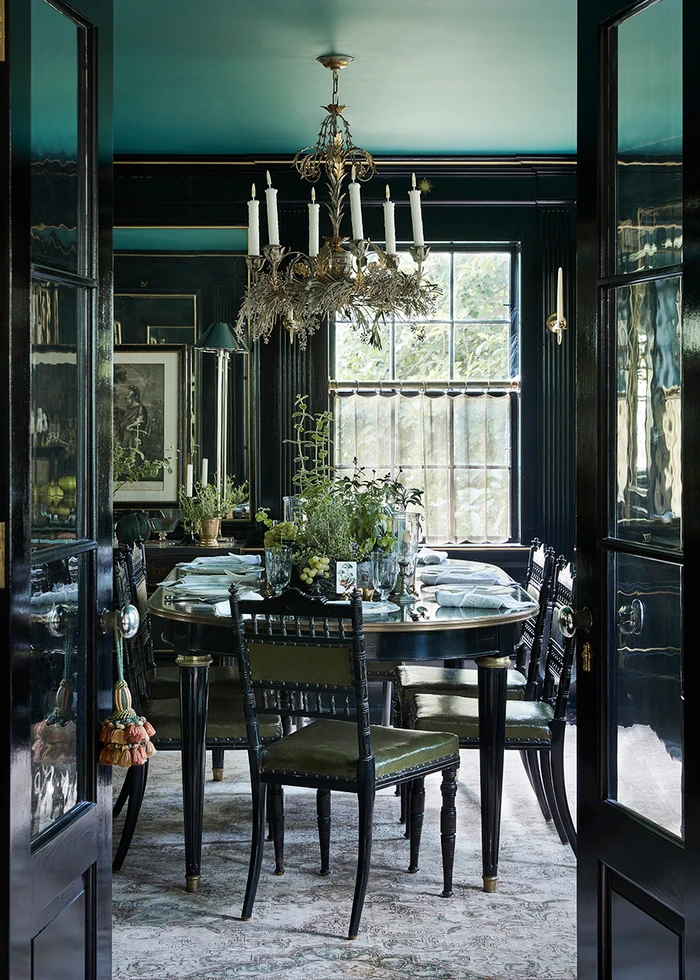 View through dark double doors into an elegant dining room styled by Sophie Strangio with a dark table, chandelier, and green accents