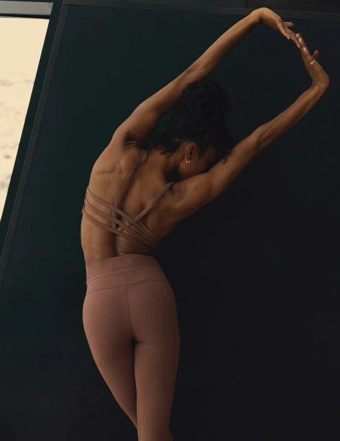 Athletic woman in sports bra and leggings stretching with arms overhead against a dark backdrop in an indoor setting, fitness theme.