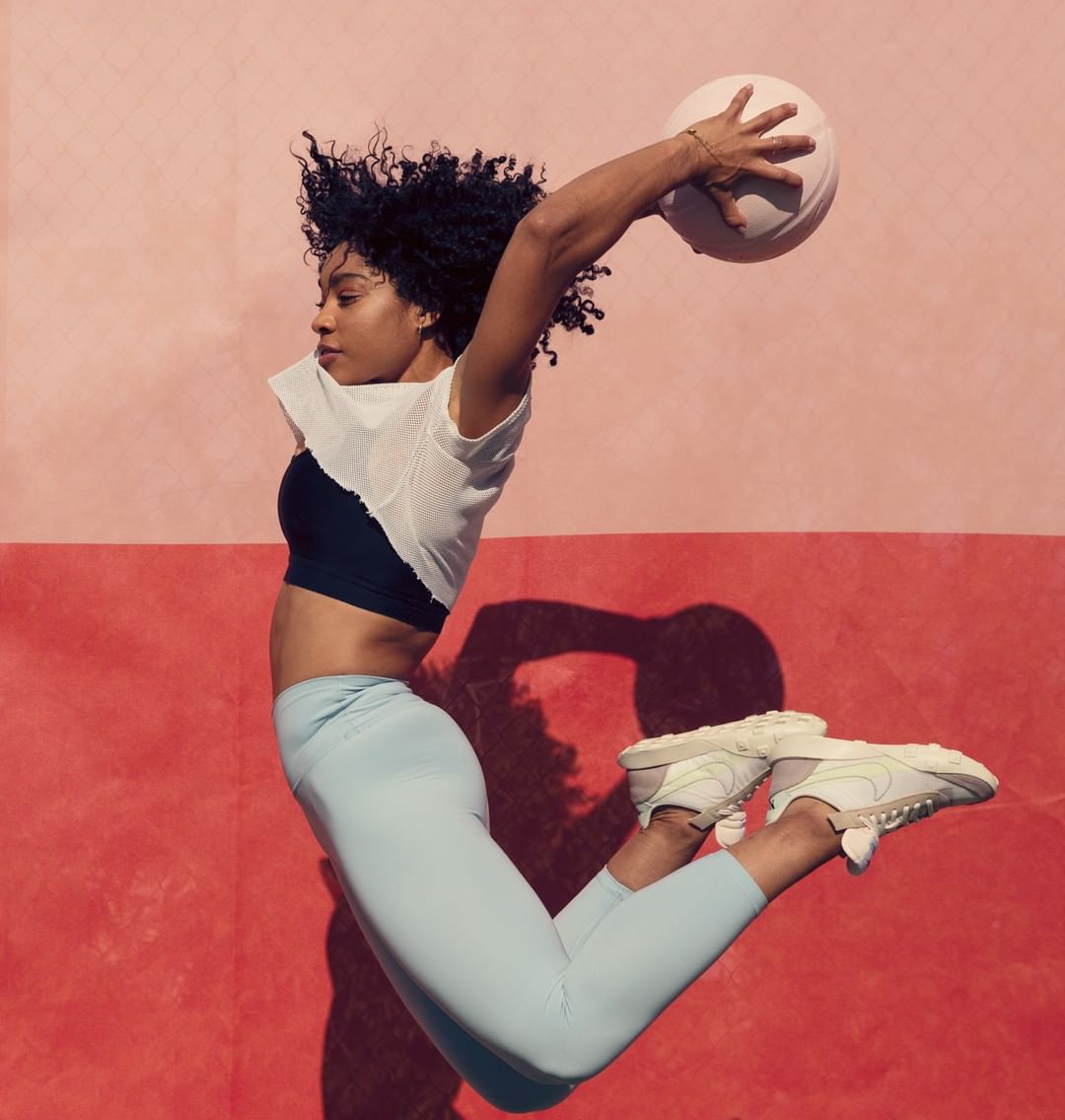 Athletic woman in sportswear leaps with a ball against a two-tone pink wall, showcasing fitness and active lifestyle photography.