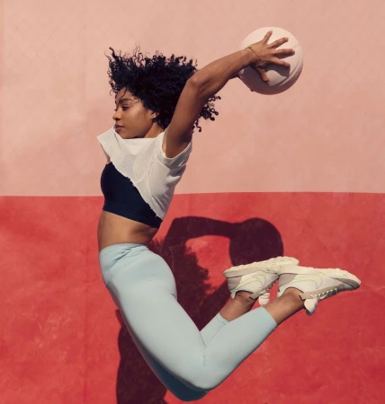 Athletic woman in sportswear leaps with a ball against a two-tone pink wall, showcasing fitness and active lifestyle photography.
