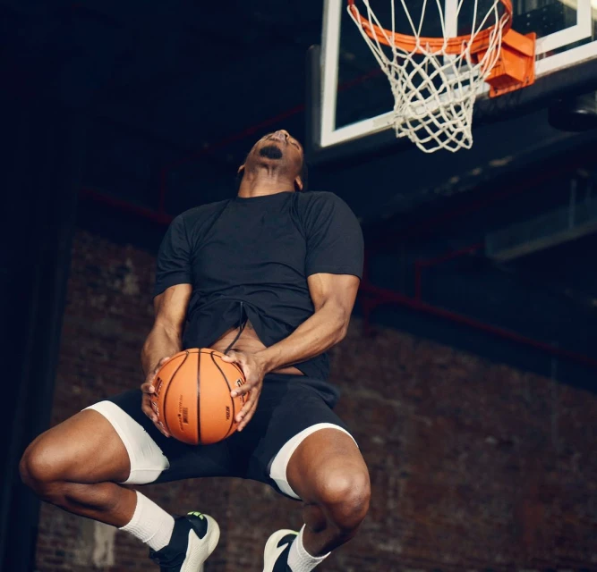 Athlete in mid-air slam dunk toward basketball hoop indoors, wearing dark sportswear, white socks and sport shoes, with the ball in hands.