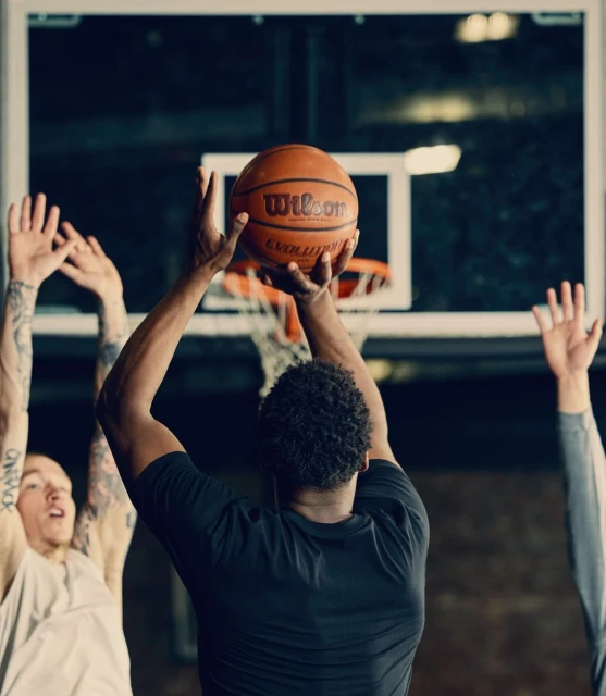 Athlete shoots a Wilson basketball at an indoor court with two other players in the background, capturing a dynamic moment in sports.