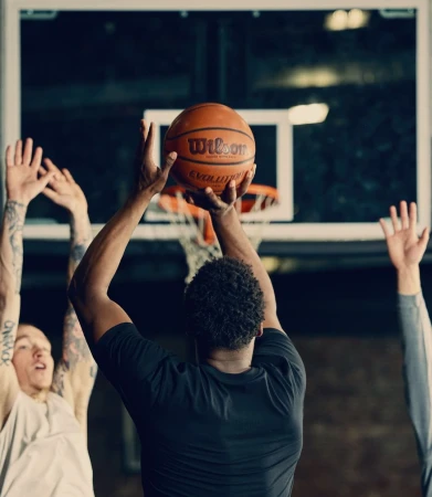 Athlete shoots a Wilson basketball at an indoor court with two other players in the background, capturing a dynamic moment in sports.