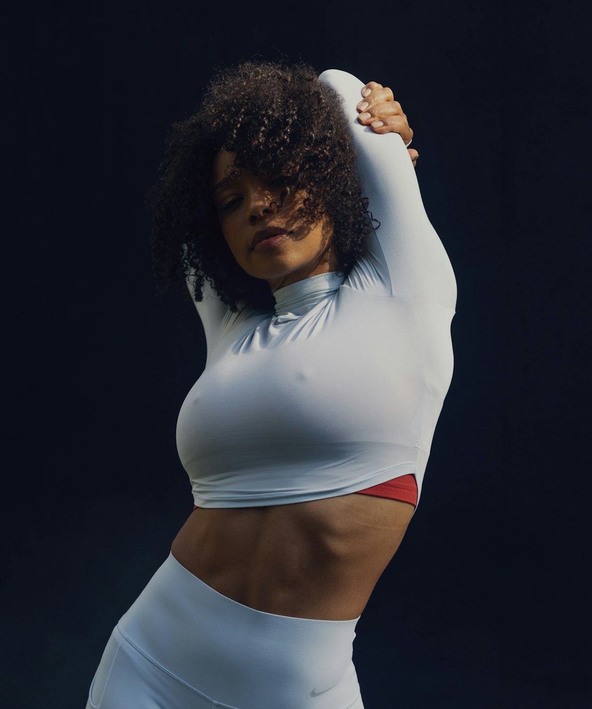 Athletic woman with curly hair in long-sleeved top and leggings stretching her arms overhead against a dark background, sportswear fashion shoot.