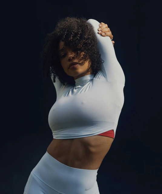 Athletic woman with curly hair in long-sleeved top and leggings stretching her arms overhead against a dark background, sportswear fashion shoot.