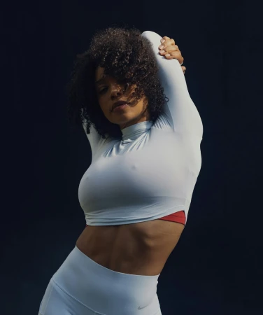 Athletic woman with curly hair in long-sleeved top and leggings stretching her arms overhead against a dark background, sportswear fashion shoot.