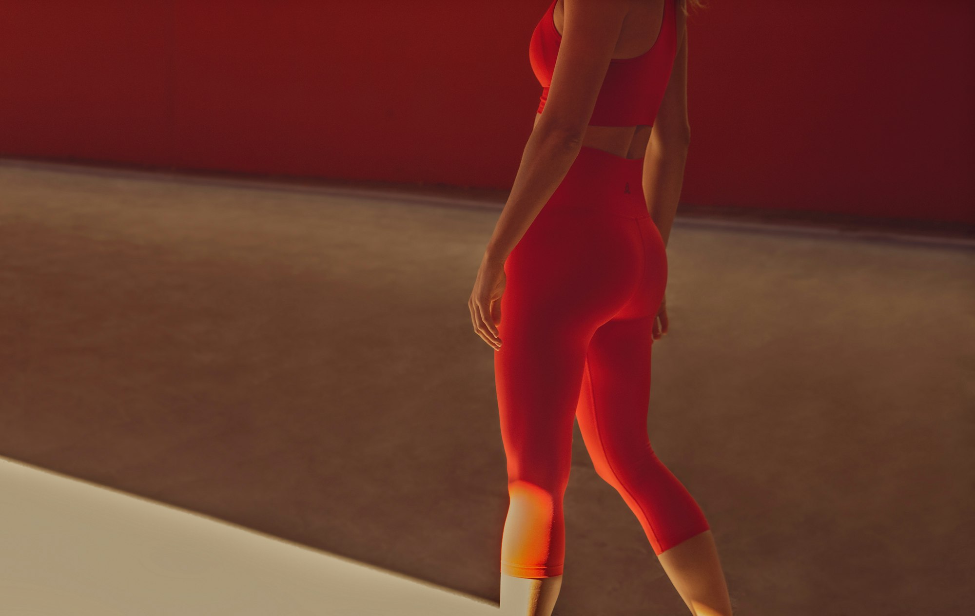 A woman is shown from the back in a red sports bra and leggings in a studio with red and tan colored walls.