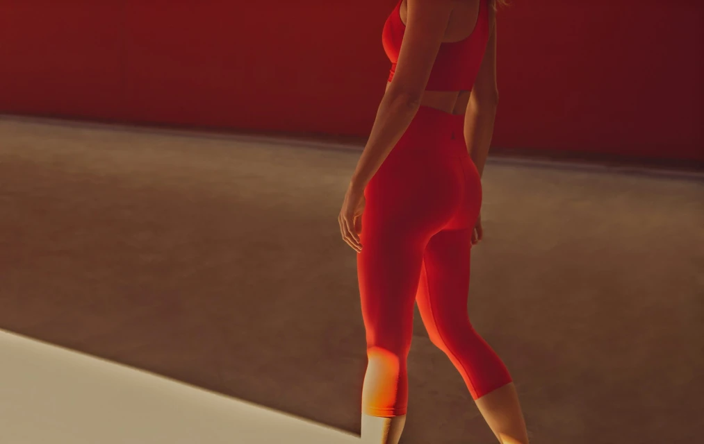 A woman is shown from the back in a red sports bra and leggings in a studio with red and tan colored walls.