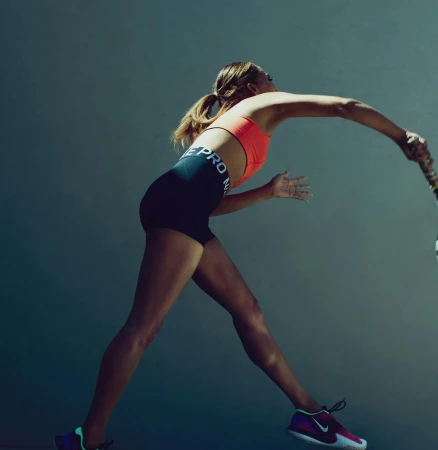 Female tennis player in action wearing an orange sports bra and black shorts swinging a racket against a grey backdrop.