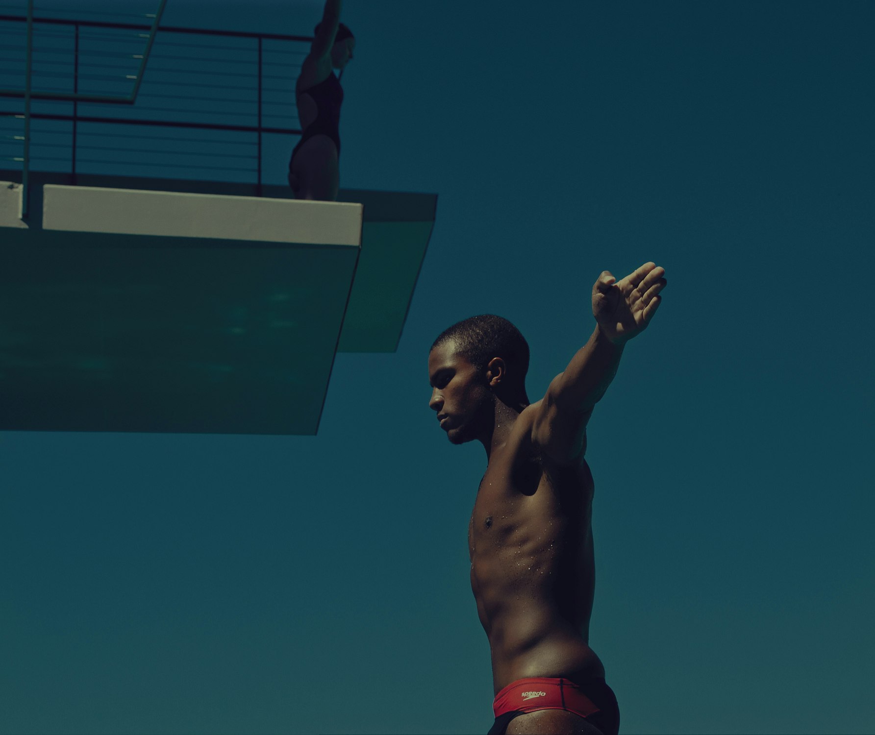 Two athletes stand poised to dive into the water at an outdoor diving platform under clear, sunny skies, showing skill and focus.