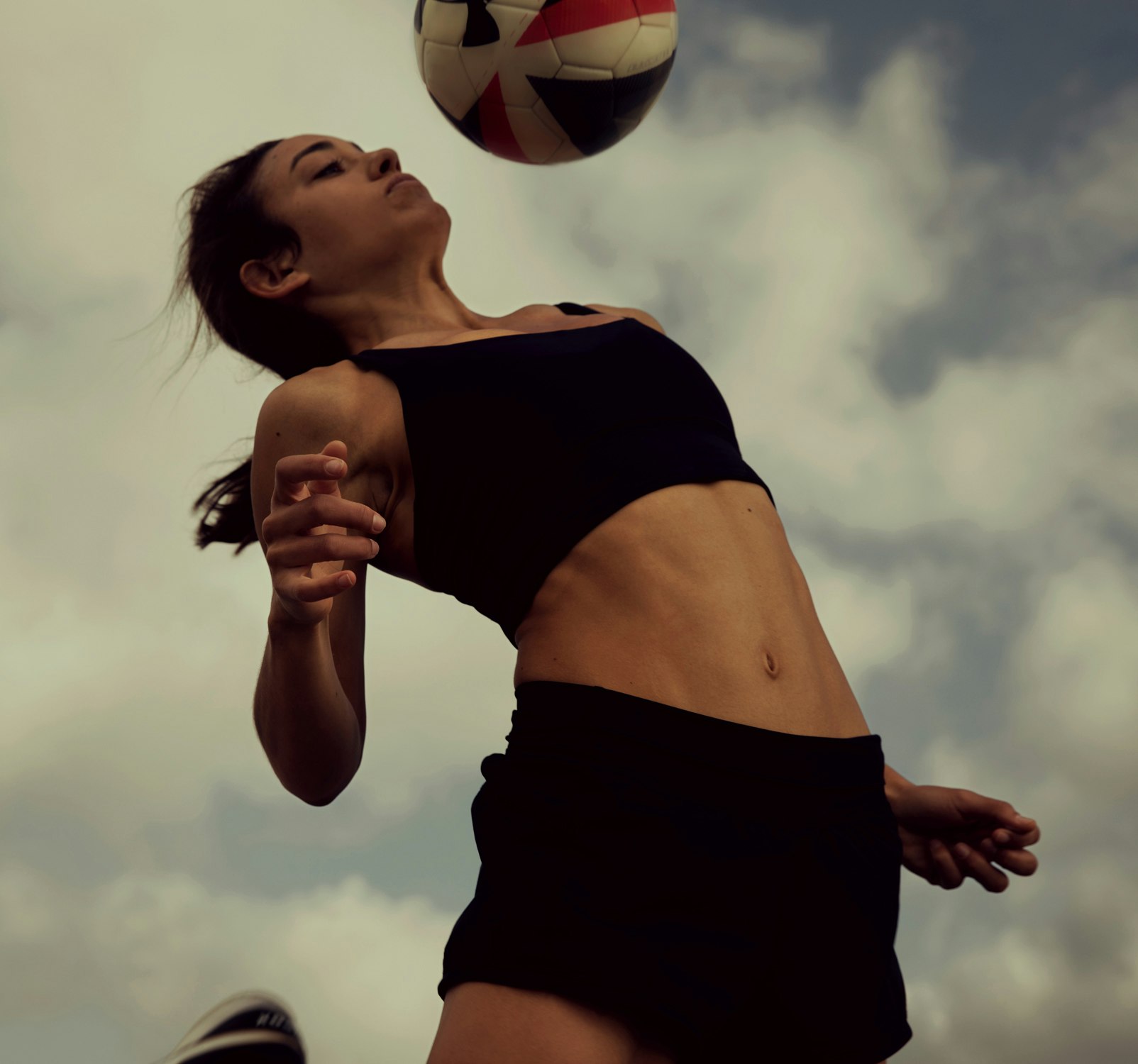 Athletic woman in black sportswear powerfully heads a multicolored soccer ball outdoors against a backdrop of cloudy skies.