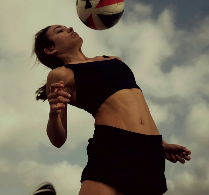 Athletic woman in black sportswear powerfully heads a multicolored soccer ball outdoors against a backdrop of cloudy skies.