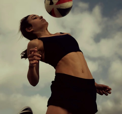 Athletic woman in black sportswear powerfully heads a multicolored soccer ball outdoors against a backdrop of cloudy skies.
