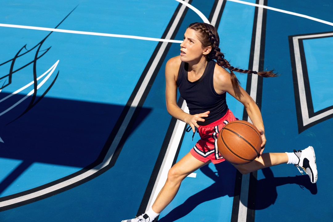 Female basketball player dribbles a basketball on an outdoor court with a bright blue painted surface and white boundary lines on a sunny day.